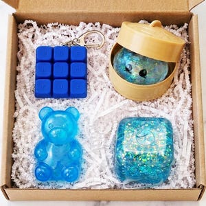 May include: A cardboard gift box containing four items: a blue square keychain with a grid of letters, a tan plastic steamer basket holding a glittery blue creature, a translucent blue bear-shaped figure, and a glittery blue square block.