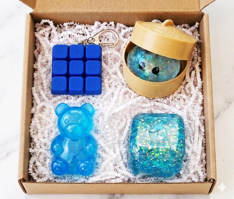 May include: A cardboard gift box containing four items: a blue square keychain with a grid of letters, a tan plastic steamer basket holding a glittery blue creature, a translucent blue bear-shaped figure, and a glittery blue square block.