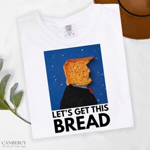 May include: White t-shirt with a graphic of a piece of toast with a bite taken out of it, wearing a suit and tie, against a blue background. The text "LET'S GET THIS BREAD" is printed below the graphic.