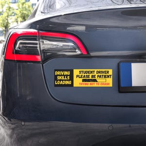 May include: A rectangular bumper sticker applied to the trunk of a dark gray car. The sticker features a black and yellow design with the text "DRIVING SKILLS LOADING" and "STUDENT DRIVER PLEASE BE PATIENT" alongside a loading bar graphic.