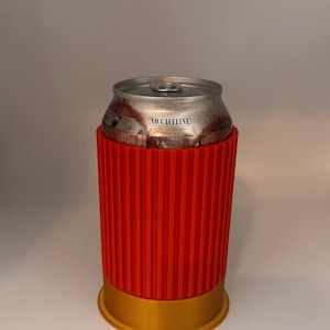 May include: A red and gold can cooler with a ribbed design, holding a silver can. The can has the text "NO CAFFEINE" printed on it. The cooler is designed to keep beverages cold.
