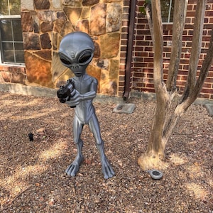 May include: A standing silver-colored alien figurine with a large head and dark, almond-shaped eyes. The figure has a slender, humanoid body and is holding a black toy pistol in its hands, pointing it forward with both arms extended outward.