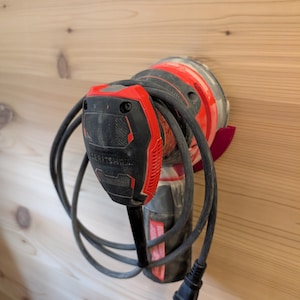 May include: A Craftsman electric sander with a red and black body and a black power cord. The sander is hanging on a light brown wooden wall. The sander has a black handle and a red base.
