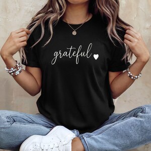 May include: Black crew neck t-shirt with the word "grateful" in white script and a small white heart. The person wearing the shirt is also wearing a gold necklace and bracelets. The t-shirt is a casual style.