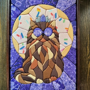 Puede incluir: Una obra de arte mural de mosaico de tela en un marco de madera oscura que muestra un gato marr&oacute;n de pelo largo con ojos de patr&oacute;n floral morado. El gato est&aacute; centrado sobre un fondo circular amarillo con chispas de colores y un borde floral.