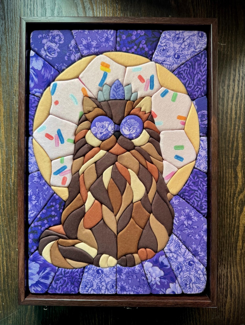 Puede incluir: Una obra de arte mural de mosaico de tela en un marco de madera oscura que muestra un gato marr&oacute;n de pelo largo con ojos de patr&oacute;n floral morado. El gato est&aacute; centrado sobre un fondo circular amarillo con chispas de colores y un borde floral.