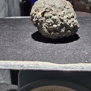 May include: A rough, textured, gray-brown rock formation. The rock is on a scale, with the dial showing a reading of 95. Several antique glass bottles are visible in the background.