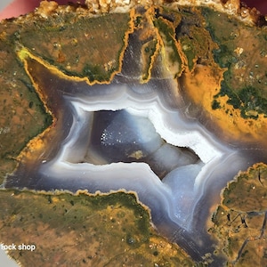 May include: A cross-section of a geode with a star-shaped center. The outer layer is a mix of brown, green, and orange hues. The center features white, blue, and gray bands. The text "Caldera rock shop" is visible.
