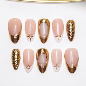 May include: A collection of ten artificial nails with a nude base and gold detailing. Designs include gold tips, open gold frames, starburst patterns, and textured elements. The nails are displayed on a white background.