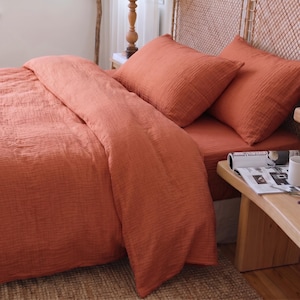 Könnte beinhalten: Ein terrakottafarbenes Bettwäsche-Set bestehend aus einem Bettbezug und zwei passenden Kissenbezügen. Der Stoff weist eine ausgeprägte, geknitterte und mehrlagige Textur auf. Die Bettwäsche ist auf einem Bett mit geflochtenem Kopfteil arrangiert.
