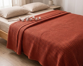 May include: A rust-colored waffle weave blanket draped over a bed with beige pillows. The blanket features a distinct grid-like texture and is accented by a decorative stem of dried white cotton bolls resting on the surface of the fabric.