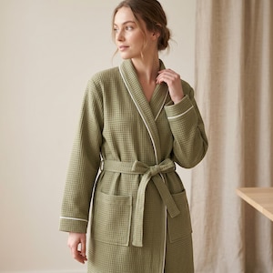 May include: An olive green waffle-knit robe featuring a shawl collar, long sleeves with white piping trim, a matching tie belt at the waist, and two large patch pockets on the front. The fabric has a distinct square grid texture throughout.