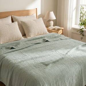 Può includere: Un letto con testiera in legno chiaro, adornato da soffici cuscini beige e una coperta verde chiaro strutturata. Un piccolo comodino in legno ospita una lampada e un libro aperto. La stanza è illuminata dalla luce naturale.