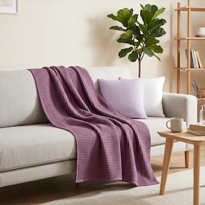 May include: A deep mauve waffle-weave throw blanket draped over a light gray sofa. The blanket features a distinct grid-like texture. Beside it, a light purple square throw pillow with a matching waffle-weave pattern rests on the sofa cushion.