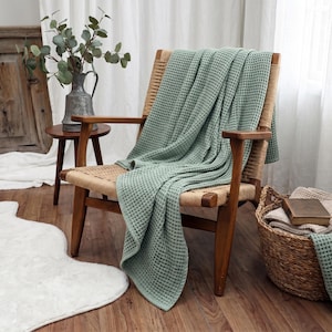 May include: A sage green waffle-weave throw blanket draped over a wooden chair with a woven seat. The blanket features a textured grid pattern and hangs over the armrest and seat of the chair, showcasing its soft fabric and consistent light green colour.
