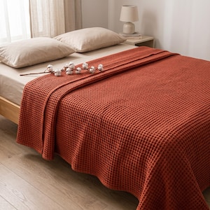 May include: A rust-colored waffle-weave blanket draped over a bed with beige pillows. A decorative stem with white cotton bolls rests on top of the textured fabric, which features a consistent grid pattern across its entire surface area.