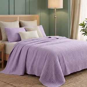 May include: A light purple textured cotton quilt draped over a bed with a cane headboard. The bedding set includes two large light purple pillows, two beige square pillows, and one small rectangular beige pillow, all arranged neatly.