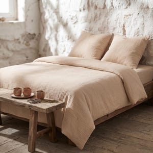 May include: A beige muslin duvet cover set with two matching square pillowcases on a wooden bed frame. A rustic wooden bench sits at the foot of the bed, holding a small plate with three ceramic cups and a sprig of dried lavender.