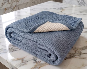Puede incluir: Una manta rectangular doblada que presenta una superficie de tela texturizada y arrugada. La capa superior es de un color azul polvoriento sólido, mientras que la parte inferior revela una tela de color crema en contraste con bordes.