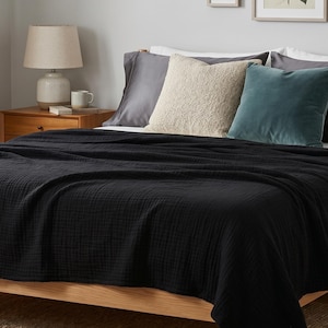 Peut inclure: Un couvre-lit noir recouvre un cadre de lit en bois, avec des oreillers gris, crème et sarcelle. Une lampe beige et une tasse sont posées sur une table de chevet en bois. La couverture est en tissu texturé.