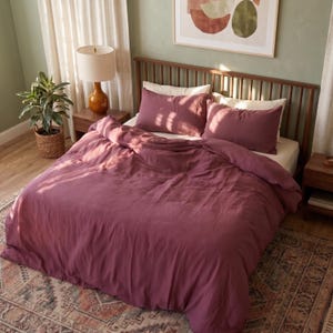 May include: A deep mauve duvet cover set featuring a textured fabric finish, including two matching rectangular pillowcases and a large comforter draped over a bed frame with a wooden headboard, creating a cohesive bedding arrangement.