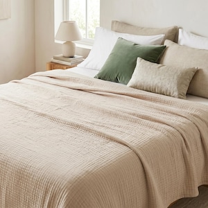 Pode incluir: Uma cama com um cobertor bege claro texturizado e várias almofadas. As almofadas incluem brancas, bege e uma almofada verde de veludo. Uma pequena lâmpada está em uma mesa de cabeceira de madeira.