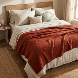 May include: A bed featuring a rust-colored textured cotton throw blanket draped over a white linen duvet. The bed includes two large white pillows, two smaller beige pillows, and one sage green square pillow, set against a light wood cane headboard.