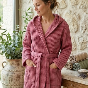 Könnte beinhalten: Ein staubrosa Waffel-Bademantel mit Kapuze, passendem Gürtel und Taschen. Der Bademantel wird von einer Person getragen, die sich in der Nähe eines Fensters und einer Steinmauer befindet. Gerollte Handtücher und eine Schale mit Badesalz stehen auf einem Tisch.