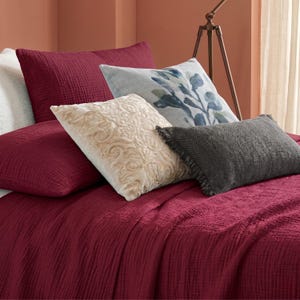 Può includere: Un set da letto composto da una trapunta stropicciata color bordeaux intenso e una federa abbinata. Il set include un cuscino quadrato color crema con ricami, un cuscino azzurro con stampa botanica e un cuscino rettangolare grigio scuro.