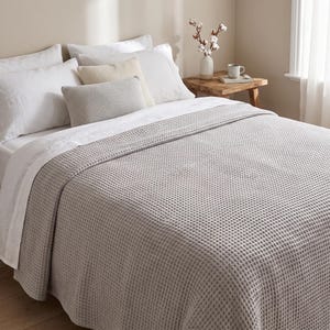 Puede incluir: Una colcha de color gris claro con tejido de gofre cubre una cama con dos almohadas blancas, una almohada cuadrada de color crema y una almohada rectangular texturizada de color gris claro. La ropa de cama está dispuesta de forma ordenada.