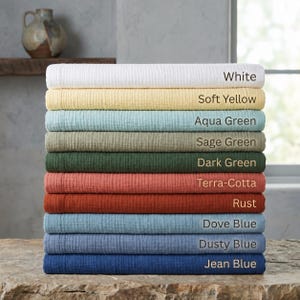 Könnte beinhalten: Ein Stapel aus zehn gefalteten Textilien in verschiedenen Farben mit den Bezeichnungen White, Soft Yellow, Aqua Green, Sage Green, Dark Green, Terra-Cotta, Rust, Dove Blue, Dusty Blue und Jean Blue. Jedes Stück hat eine texturierte Oberfläche.