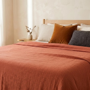 May include: A terracotta-colored crinkled cotton bedspread covers a bed featuring a light wooden headboard. The bed is styled with a beige linen pillow, a burnt orange velvet square pillow, and a dark gray textured rectangular throw pillow.