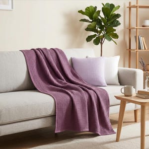 May include: A deep mauve waffle-weave throw blanket draped over a light gray sofa. A square light purple waffle-weave throw pillow sits on the sofa next to the blanket. The fabric features a distinct grid-like texture throughout the surface.