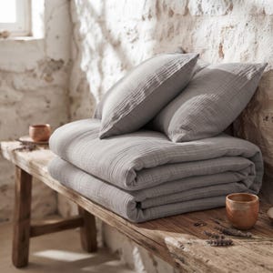 May include: A folded light gray muslin bedding set consisting of a thick, textured quilt and two matching rectangular pillowcases. The fabric features a subtle, crinkled, multi-layered weave pattern throughout the entire surface of the bedding.