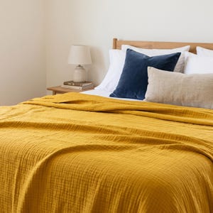 Può includere: Un copriletto giallo senape copre un letto con lenzuola e cuscini bianchi. Un cuscino di velluto blu navy e un cuscino di lino beige poggiano sulla testiera. Una piccola lampada e libri sono su un comodino di legno.