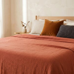 May include: A bed with a terracotta-coloured, textured bedspread. The bed has a wooden headboard and several pillows in beige, brown, and grey. A small vase with dried flowers sits on a wooden side table.