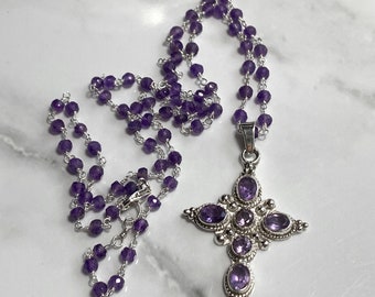 May include: A silver necklace with a cross pendant featuring amethyst gemstones. The necklace is made of a silver chain with amethyst beads.