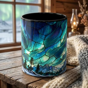 May include: A ceramic mug with a stained-glass design depicting a mountain landscape under a vibrant aurora borealis. The mug features shades of blue, teal, and black, with a black interior and a detailed forest scene at the base.