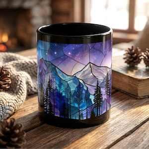 May include: Black ceramic mug featuring a stained-glass design of a mountain range and forest under a starry night sky. The design incorporates shades of blue, purple, and white. The mug is cylindrical and sits on a wooden surface.
