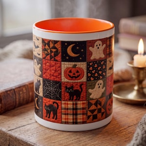 May include: A cylindrical ceramic mug with a bright orange interior and a white exterior featuring a patchwork quilt pattern. The design includes squares with black cats, ghosts, jack-o'-lanterns, spiderwebs, crescent moons, and plaid fabric.