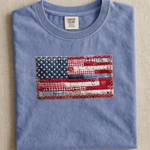 May include: A pale blue t-shirt with a sewn-on American flag design. The flag is a patchwork of red, white, and blue fabrics. The shirt has a crew neck and a label that reads "Comfort Colors".