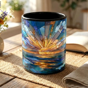 May include: A cylindrical ceramic mug with a black interior and a stained-glass design depicting a sunset over the ocean. The design features shades of blue, orange, and yellow. The mug sits on a wooden table.