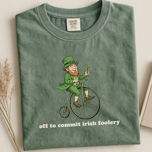 May include: Green t-shirt featuring a cartoon leprechaun riding a penny-farthing bicycle and holding a four-leaf clover. The text "off to commit irish foolery" is printed below the illustration. The shirt has a crew neck and a tag.