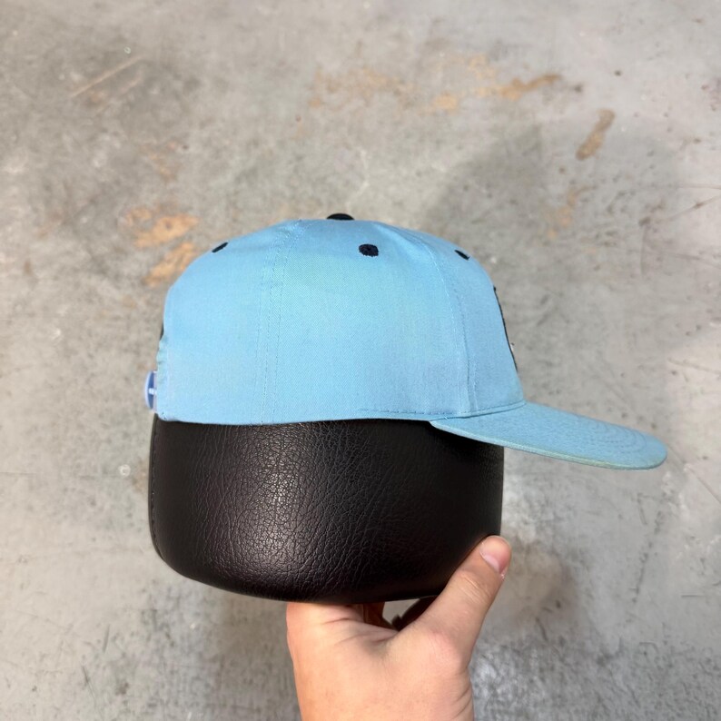May include: A light blue baseball cap with a black brim. The cap has a curved bill and a structured crown. The cap is being held by a hand. The cap has a classic, sporty design.