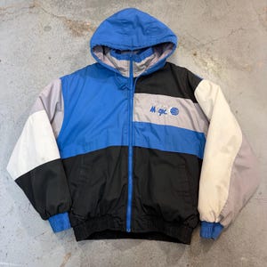 May include: A vintage hooded jacket in a colour block design of blue, black, white, and grey. The jacket has a full zip, elastic cuffs, and the word "Magic" with a basketball graphic on the chest. It features a blue hood and a grey lining.
