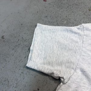 Puede incluir: Primer plano de una manga de camiseta gris claro con un dobladillo enrollado. La tela tiene una textura jaspeada, que muestra una mezcla de fibras grises claras y oscuras. La manga est&aacute; en &aacute;ngulo, mostrando la costura y el dobladillo enrollado.