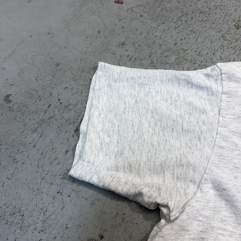 Puede incluir: Primer plano de una manga de camiseta gris claro con un dobladillo enrollado. La tela tiene una textura jaspeada, que muestra una mezcla de fibras grises claras y oscuras. La manga est&aacute; en &aacute;ngulo, mostrando la costura y el dobladillo enrollado.