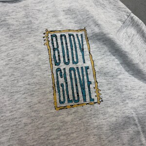 Puede incluir: Una sudadera gris con un gr&aacute;fico rectangular con las palabras "BODY GLOVE" en un dise&ntilde;o turquesa y amarillo. La sudadera est&aacute; hecha de una tela suave y texturizada.