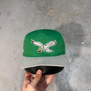 Puede incluir: Gorra de béisbol verde y gris con un águila blanca sosteniendo un balón de fútbol. La gorra tiene una visera curva y una corona estructurada. Un accesorio de temática deportiva.