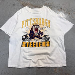 May include: A white short-sleeve t-shirt featuring a graphic of the Tasmanian Devil character centered between two Pittsburgh Steelers football helmets. The text PITTSBURGH appears in yellow above the character, and STEELERS appears below.